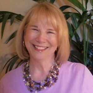 smiling woman, looking forward, short blond hair, wearing a lavendar blouse and beaded necklace