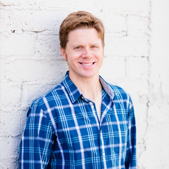 Person wearing plaid blue and white collard shirt, looking directly forward, smiling, and leaning against a white brick building.