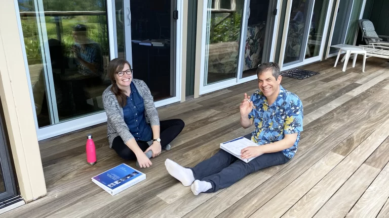 Two adults sitting outside on a wood deck, course materials spread around them, both smiling.
