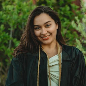 Adult, outdoors, wearing green graduation robe, smiling, looking directly ahead.