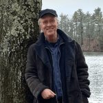 Photograph of George Kinder standing outdoors, wearing casual jacket and hat, smiling and looking directly forward.