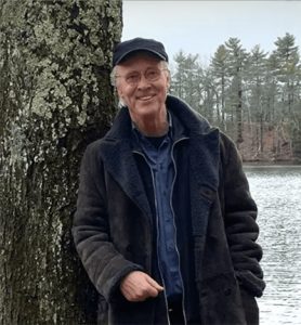 Photograph of George Kinder standing outdoors, wearing casual jacket and hat, smiling and looking directly forward.