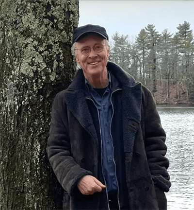Photograph of George Kinder standing outdoors, wearing casual jacket and hat, smiling and looking directly forward.