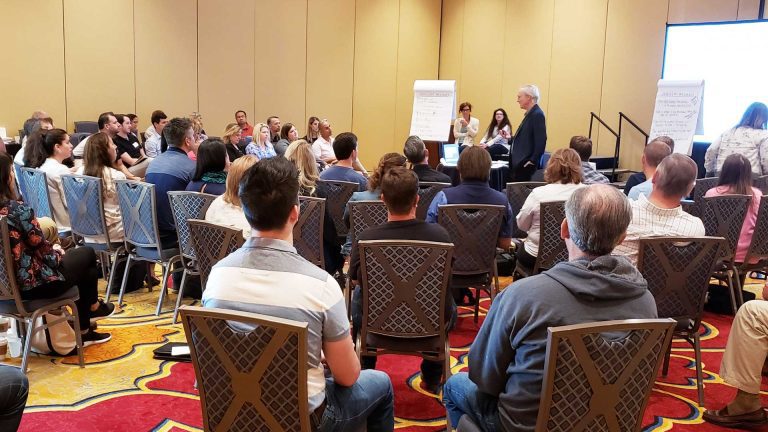 Large conference room filled with many adults sitting and facing the speaker who is actively speaking to the attendees.