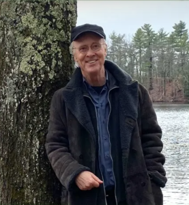 Photograph of George Kinder, standing next to tree, wearing casual jacket and hat