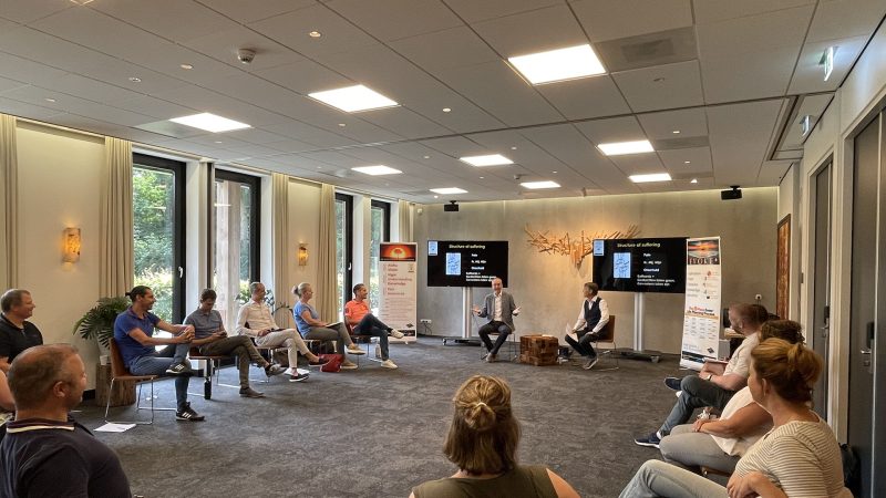 home-hero-kinder-institute-1920×1080 Group of 14 adults sitting in a circle indoors, wearing casual clothes. Two of the adults are leading the others in a lively discussion.